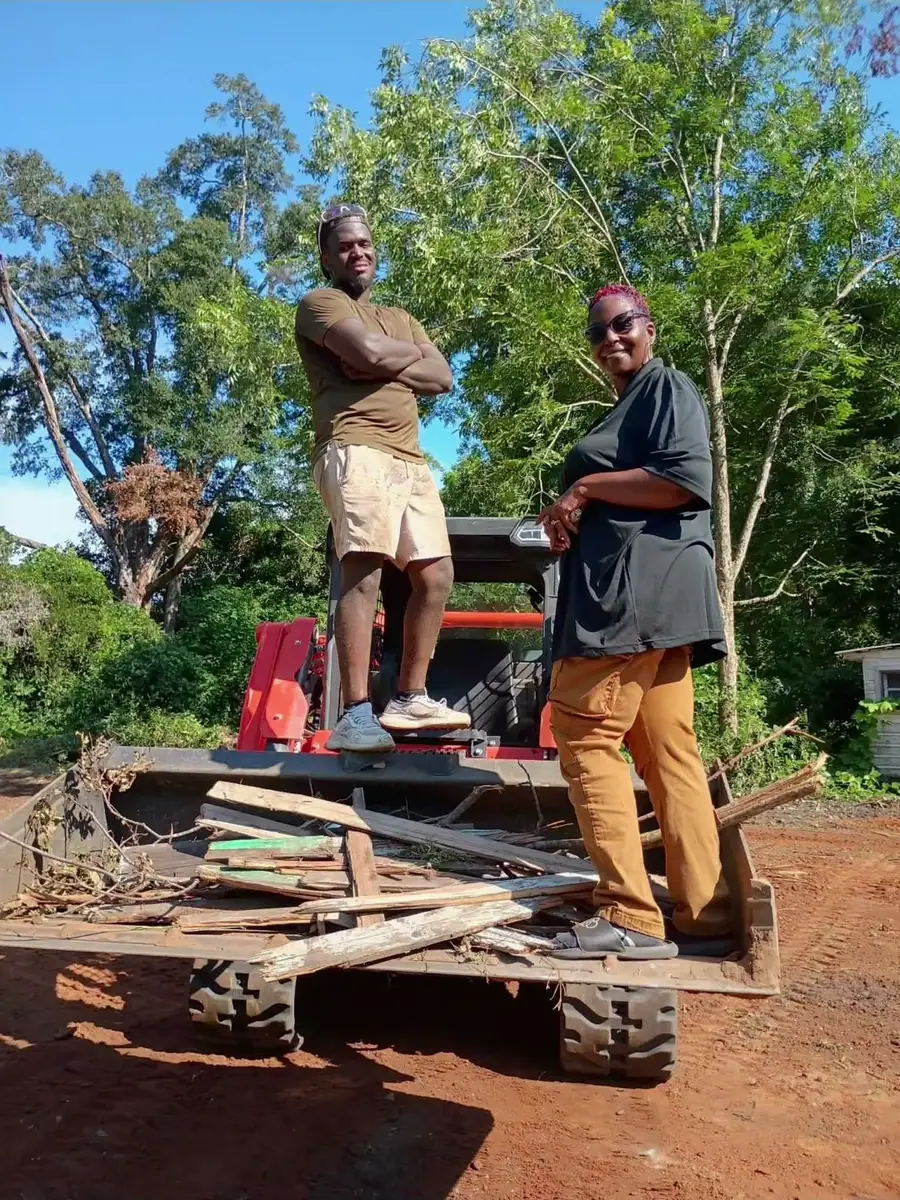 Two people standing in the bucket of a red track loader on a cleared red-dirt lot
