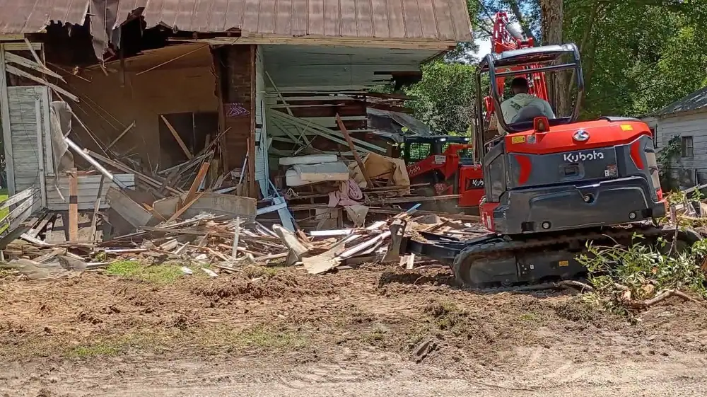 Kubota mini excavator demolishing an old frame house with a rusted metal roof