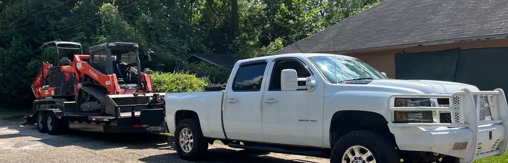 White crew-cab pickup towing a trailer loaded with a Kubota track loader and a mini excavator